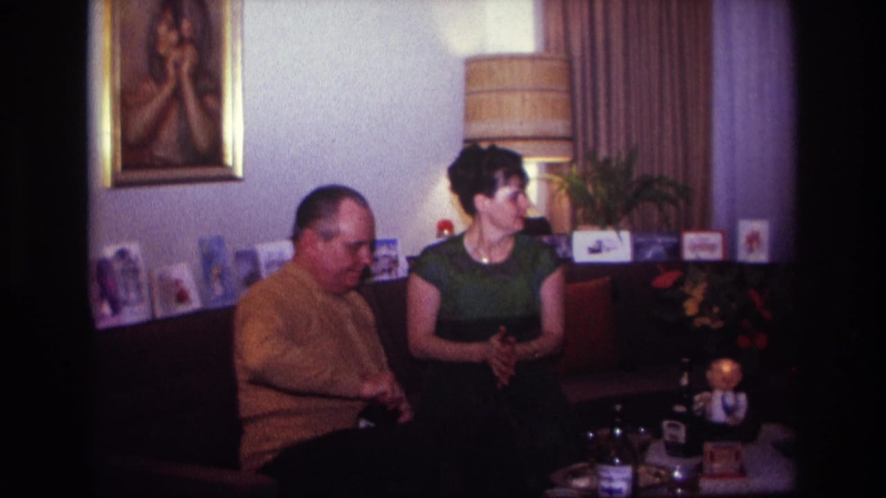 1967 Rockaway Beach home-movie frame of a man in a mustard sweater and a woman in green seated in a decorated living room.