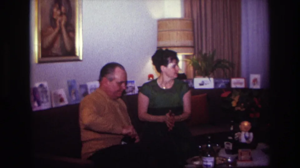 1967 Rockaway Beach home-movie frame of a man in a mustard sweater and a woman in green seated in a decorated living room.