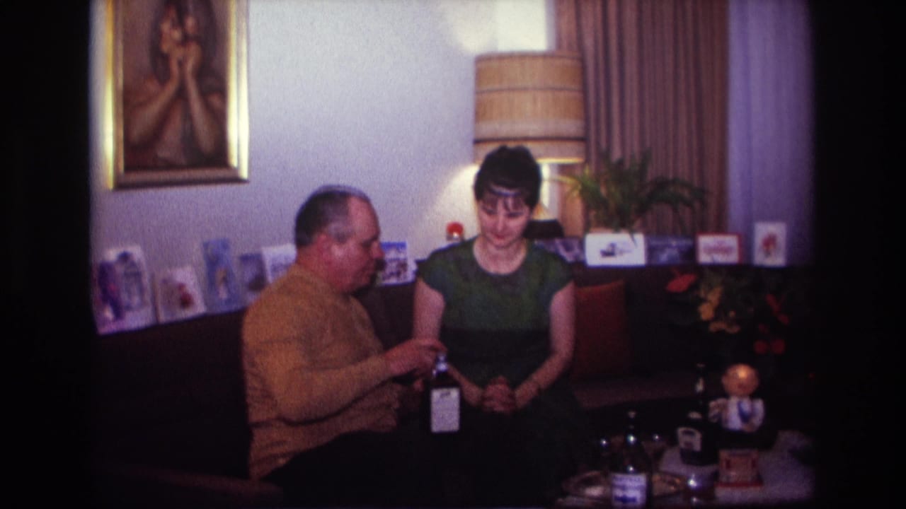 Seated man holds a bottle while the woman looks down beside him on the couch.