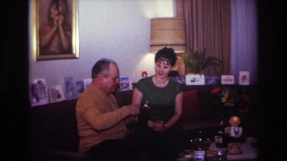 1967 Rockaway Beach interior frame with a seated man working at a bottle and a woman watching in a softly lit room.