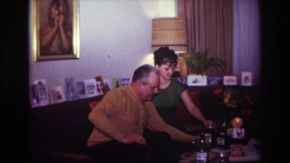 1967 Rockaway Beach home-movie frame of a man placing or reaching with a bottle near a coffee table as a woman sits nearby.