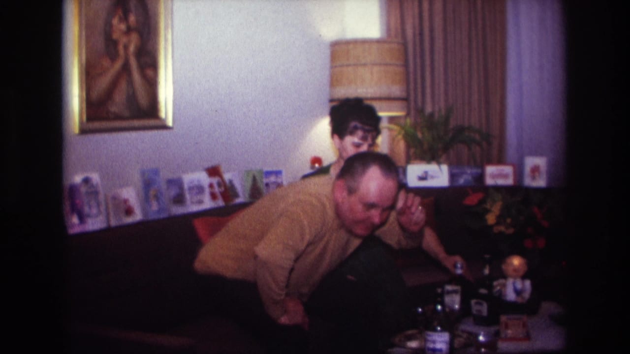 1967 Rockaway Beach home-movie frame showing a man leaning toward the camera in a living room with a seated woman, cards, bottles, and wall art.