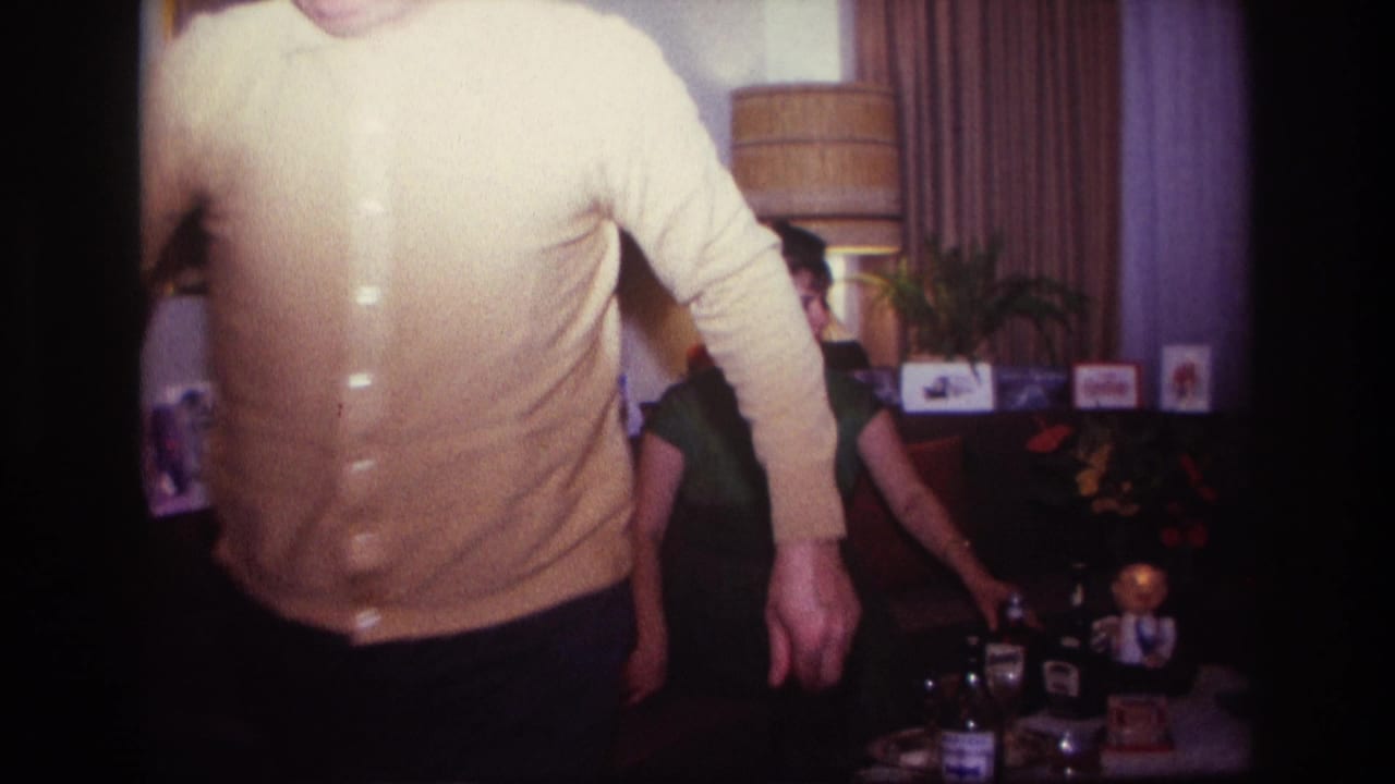 Man in yellow cardigan passes close to the camera as a woman sits near a table with bottles.