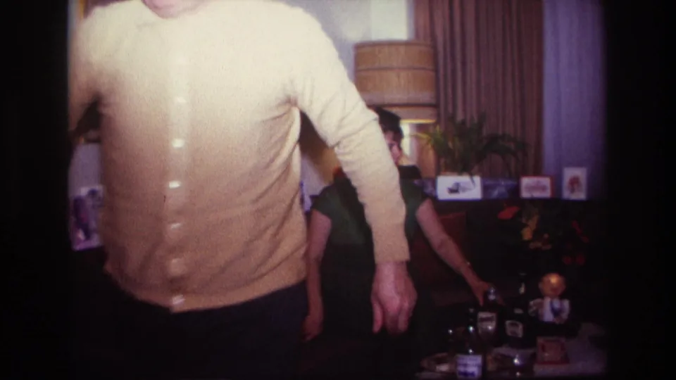 1967 Rockaway Beach home-movie frame with the man moving close to the lens and the woman seated near liquor bottles in the living room.
