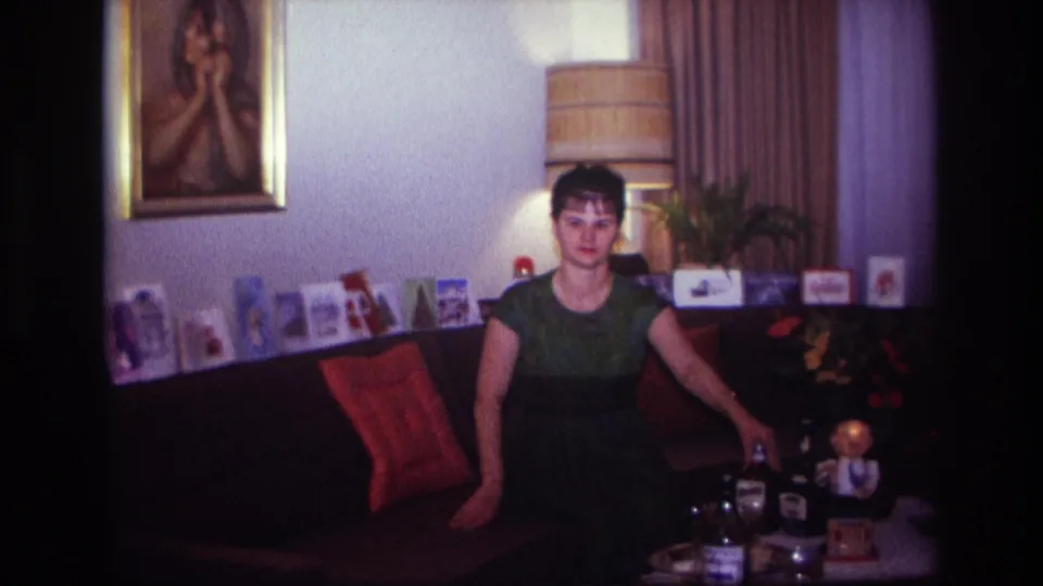 1967 Rockaway Beach home-movie frame showing a woman in a green dress seated on a couch beside bottles, glasses, cards, and a lamp.