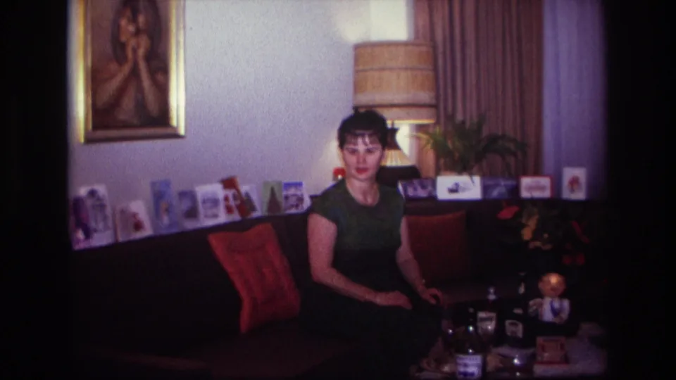 1967 Rockaway Beach home-movie frame of a woman seated in a private living room with liquor bottles, glasses, greeting cards, lamp, and wall art.