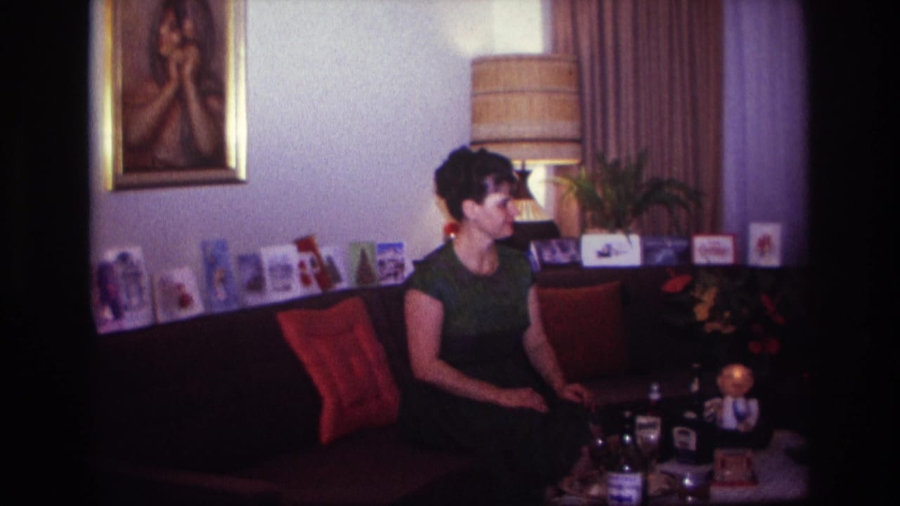 1967 Rockaway Beach home-movie frame of a woman seated on a sofa in a warmly lit living room with photos, cushions, and a table lamp.