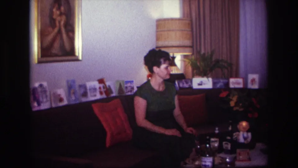 1967 Rockaway Beach home-movie frame of a woman seated on a sofa in a warmly lit living room with photos, cushions, and a table lamp.