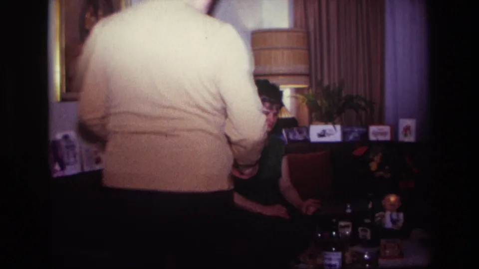 1967 Rockaway Beach living-room frame with a man in a cream sweater entering near a seated woman on the sofa.