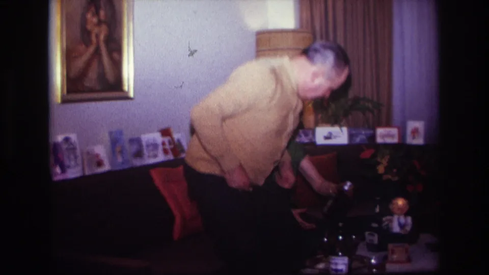 1967 Rockaway Beach home-movie frame showing a man bending toward a seated woman in a living room with drinks on a table.