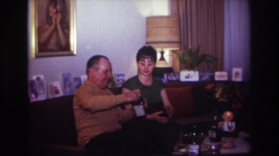 1967 Rockaway Beach home-movie frame of two adults seated together on a sofa, with the man holding a bottle near a small table.