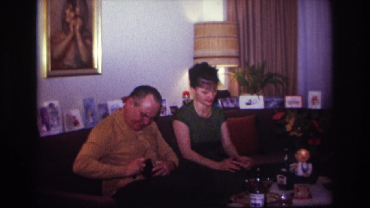 1967 Rockaway Beach home-movie frame showing two adults on a couch, with the man handling a dark bottle and the woman seated beside holiday cards.