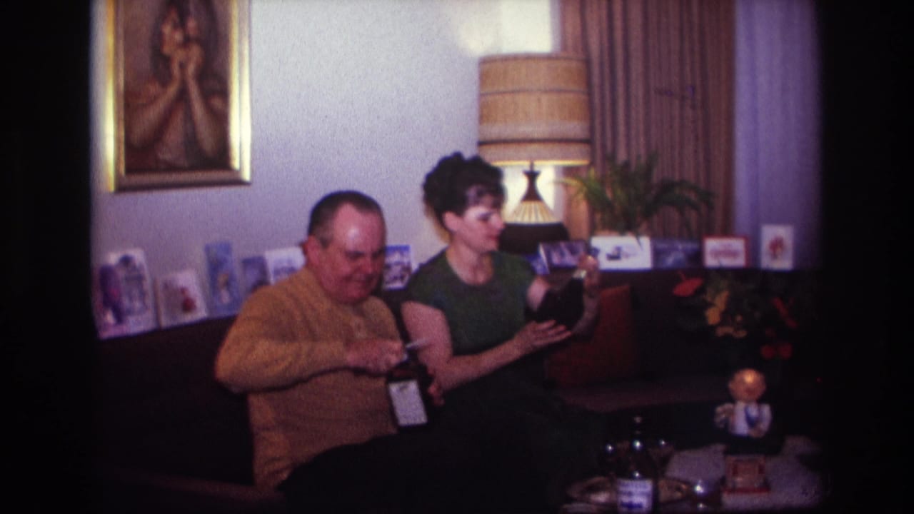 Man works with a bottle while woman holds a dark object on a couch in a living room.