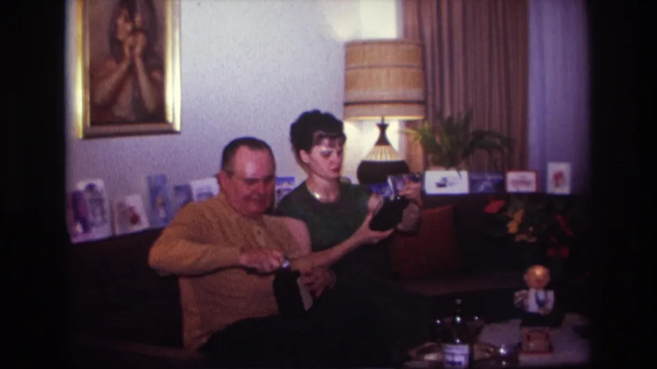 1967 Rockaway Beach home-movie frame showing a woman presenting a dark bottle-shaped item as a man beside her works with an opener.