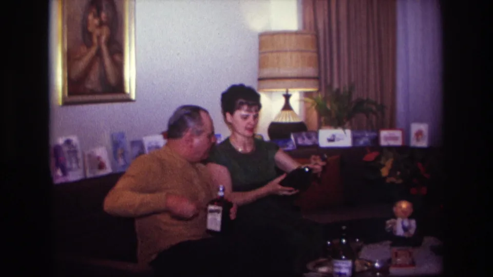 1967 Rockaway Beach home-movie frame showing the man turning toward the woman as she holds a dark bottle-shaped object in the living room.
