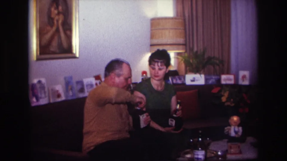 A 1960s home-movie frame shows the couple coordinating drinks on the couch in a private living room.
