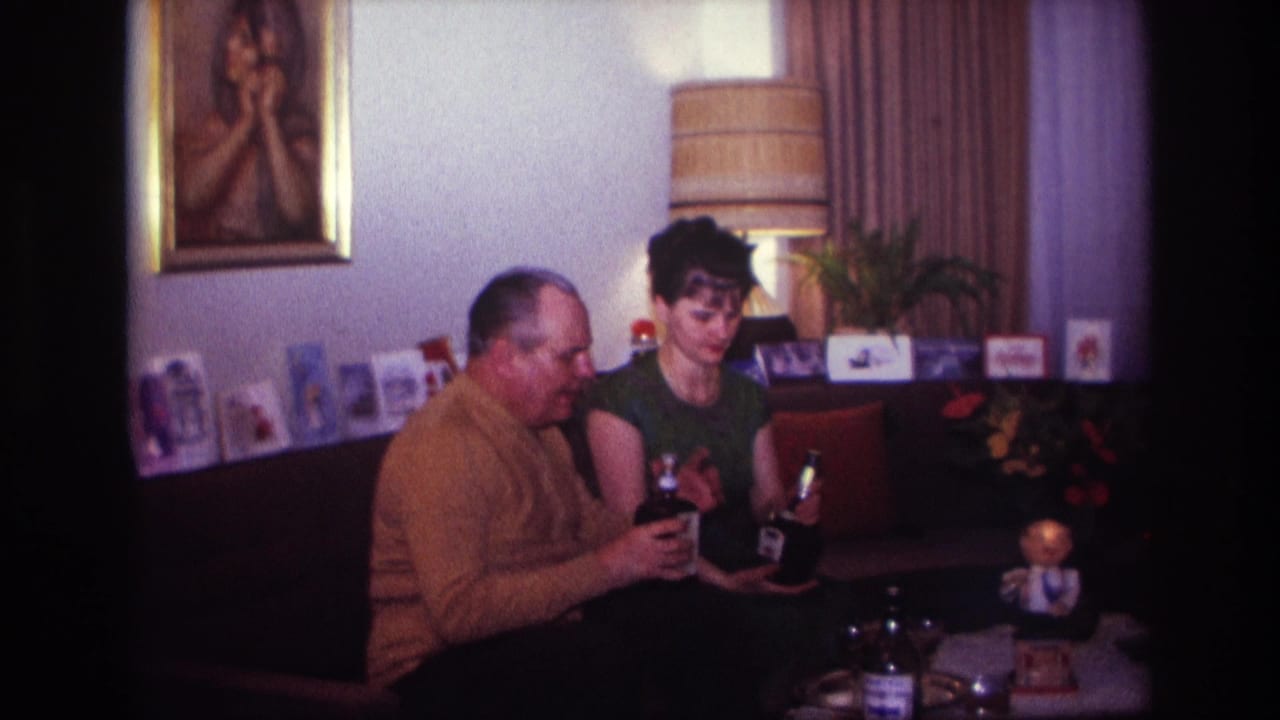 In a 1967 Rockaway Beach home movie frame, two adults sit close together on a dark couch with drinks, cards, a lamp, and wall art behind them.