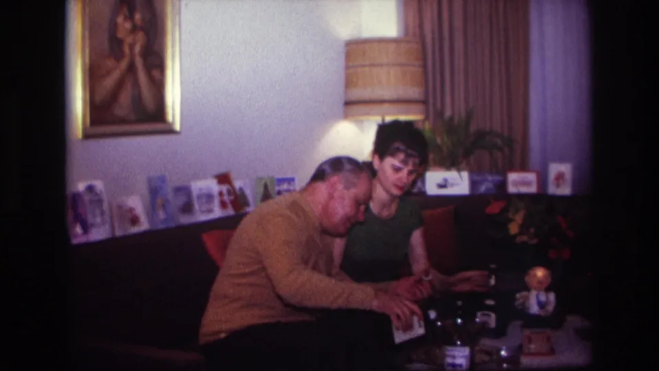 In the living room, the man bends forward over the table and the woman looks down, with cards, a framed portrait, lamp, and drinks around them.