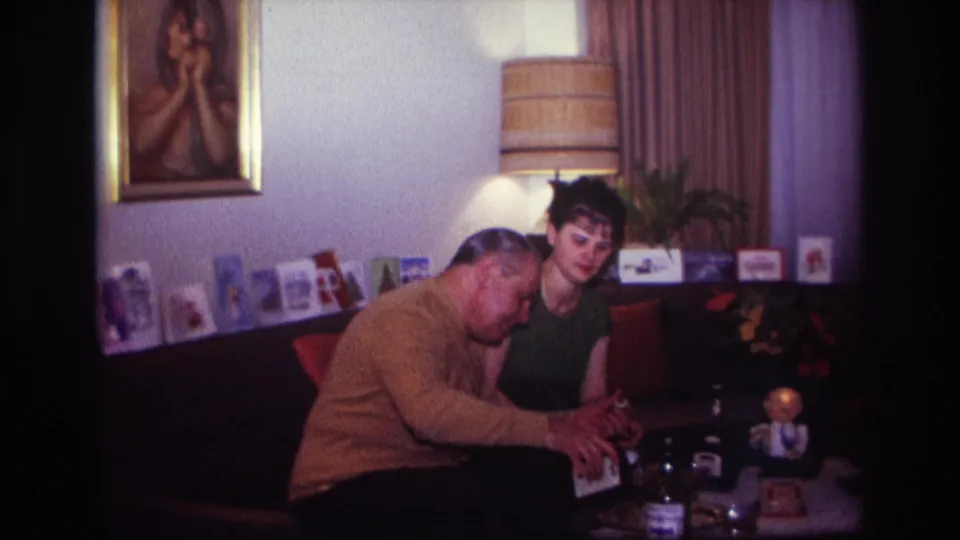 The pair remain seated close together in the Rockaway Beach living room, surrounded by drinks, a table lamp, greeting cards, curtains, and wall art.