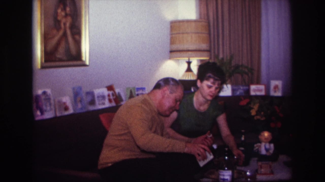 In a 1967 Rockaway Beach home-movie frame, a man in a mustard sweater leans over table drinks beside a woman in a green top.