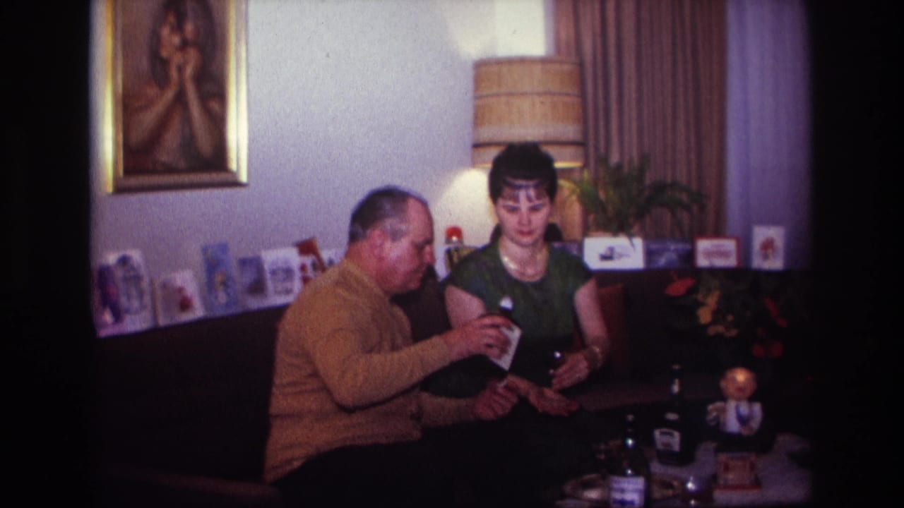 Man holds a drink container near a seated woman in a living room with cards and a lamp behind them.