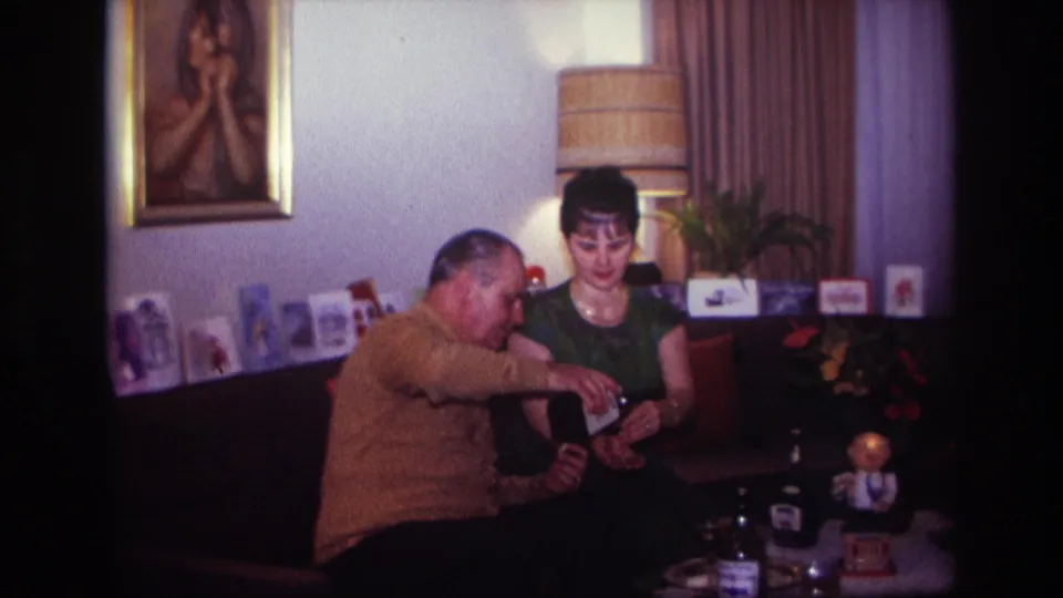 A man pours from a bottle as a woman in a green top watches the glass in a private 1967 Rockaway Beach living room.