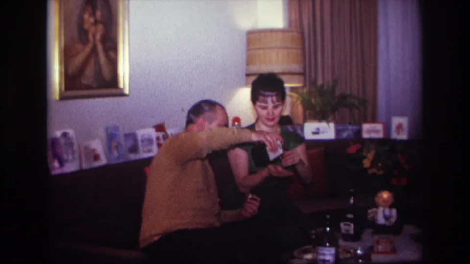 The man extends a bottle toward the woman's glass in a cozy living room, with cards, lamp, painting, and table objects visible.