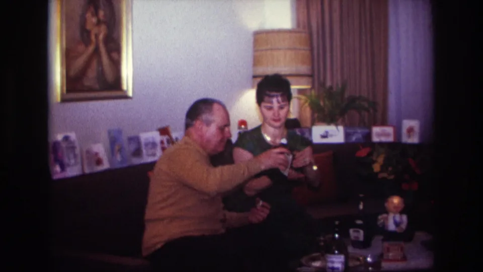 1967 Rockaway Beach home-movie frame of two adults on a couch as the man handles a liquor bottle and the woman holds a glass.