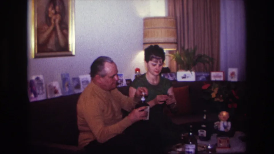 1967 Rockaway Beach living-room frame with a man handling a liquor bottle and a woman seated beside him with a glass or small item.