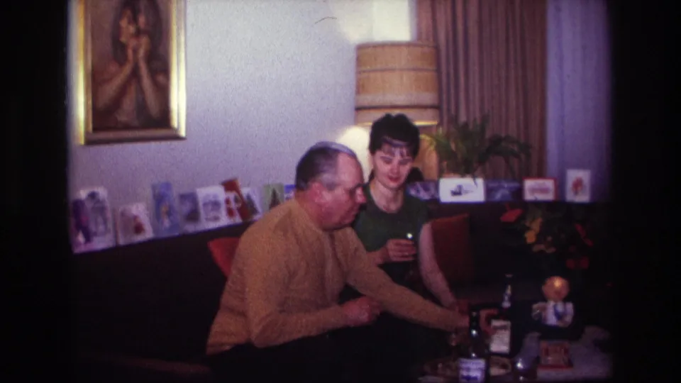 1967 Rockaway Beach living-room frame of a man reaching toward table bottles as a woman remains seated with a glass.