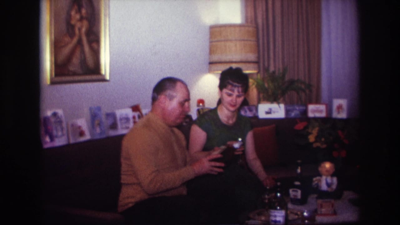 In a 1967 Rockaway Beach home movie frame, a man in a mustard sweater and a woman in green sit together with glasses near a low table.