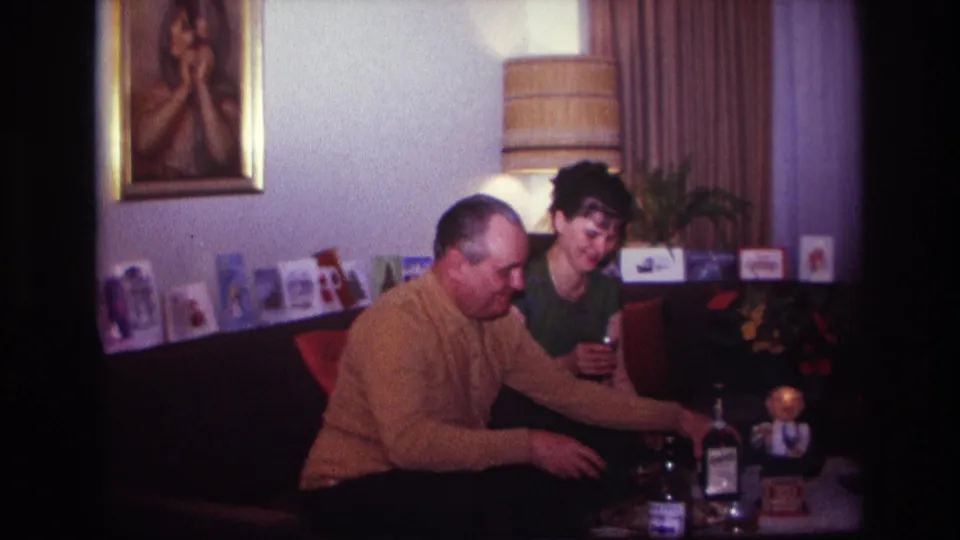 In the private living room, the man leans toward the table with bottles while the woman remains seated holding a drink.