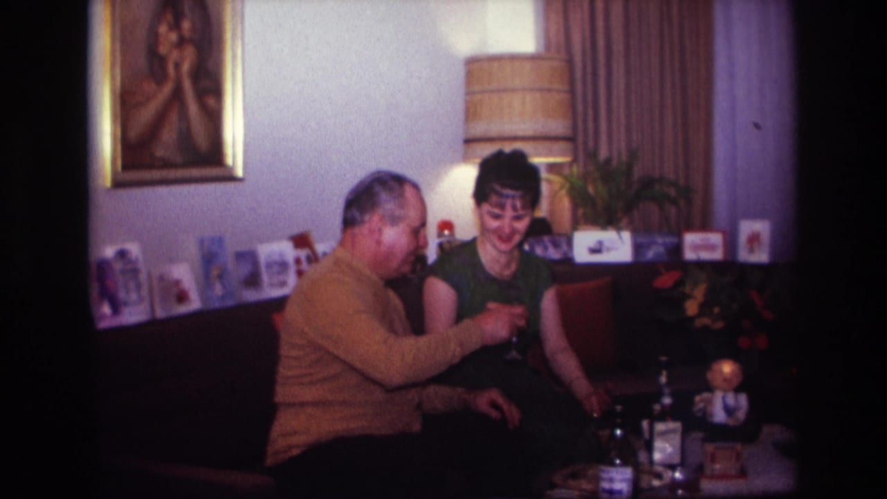 1967 Rockaway Beach home-movie frame of a seated couple in a living room, with glasses, bottles, cards, a lamp, and a framed painting visible.