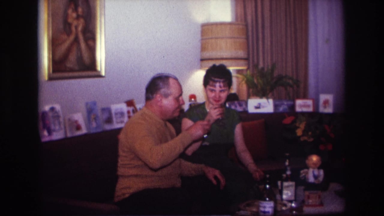 The man in a mustard sweater raises his glass toward the woman in a green dress inside the Rockaway Beach living-room scene.