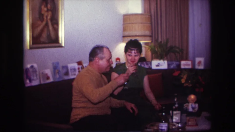 The man in a mustard sweater raises his glass toward the woman in a green dress inside the Rockaway Beach living-room scene.