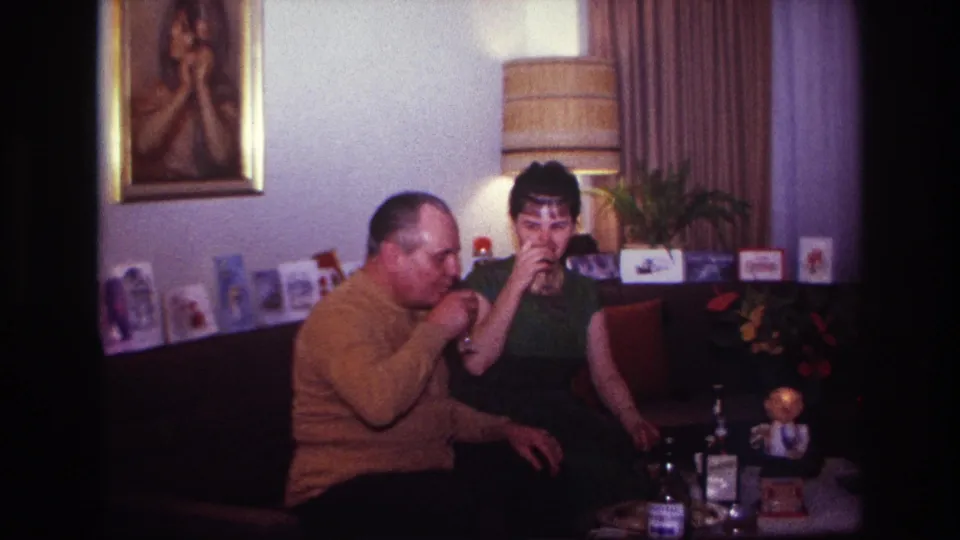1967 home-movie frame showing the couple drinking together on a couch, surrounded by living-room decor and a small table arrangement.