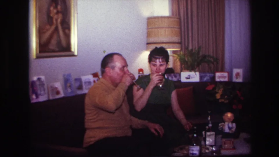 The couple drinks together in the Rockaway Beach living room, with cards, a lamp, curtains, bottles, and a small figurine in view.