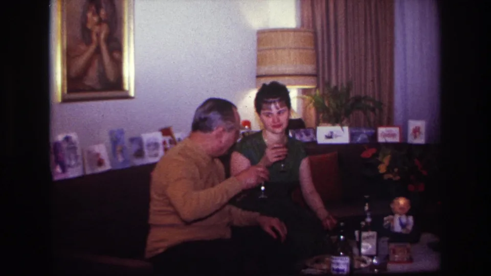 1967 Rockaway Beach home-movie frame showing a couple seated close together, wine glasses raised, with greeting cards and living-room decor visible.