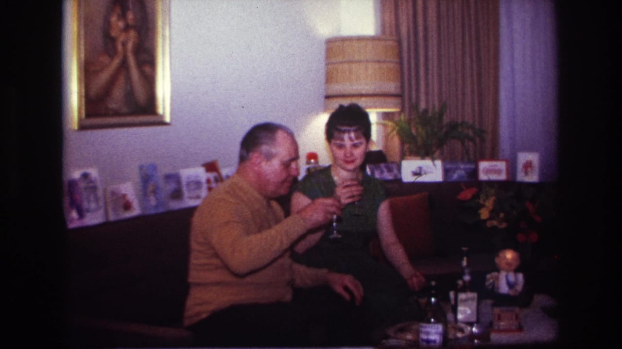 In a 1967 Rockaway Beach home interior, a man in a mustard sweater and a woman in a dark dress sit together with drinks.
