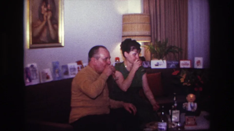 The man and woman sip their drinks in a Rockaway Beach living room, with a striped lamp and greeting cards behind them.