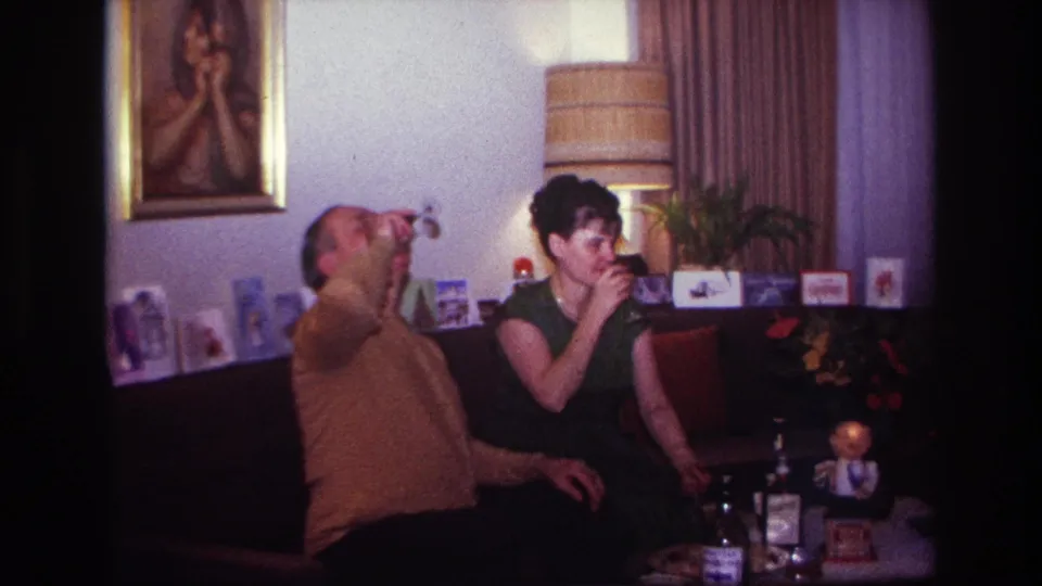 A man raises his glass high as the woman continues sipping, framed by 1960s home decor and greeting cards.