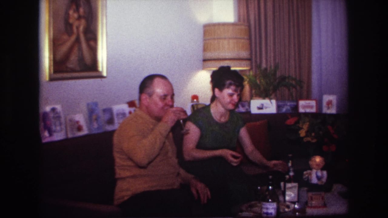1967 Rockaway Beach home-movie frame of a man and woman seated on a couch with drinks, bottles, greeting cards, and a lamp behind them.