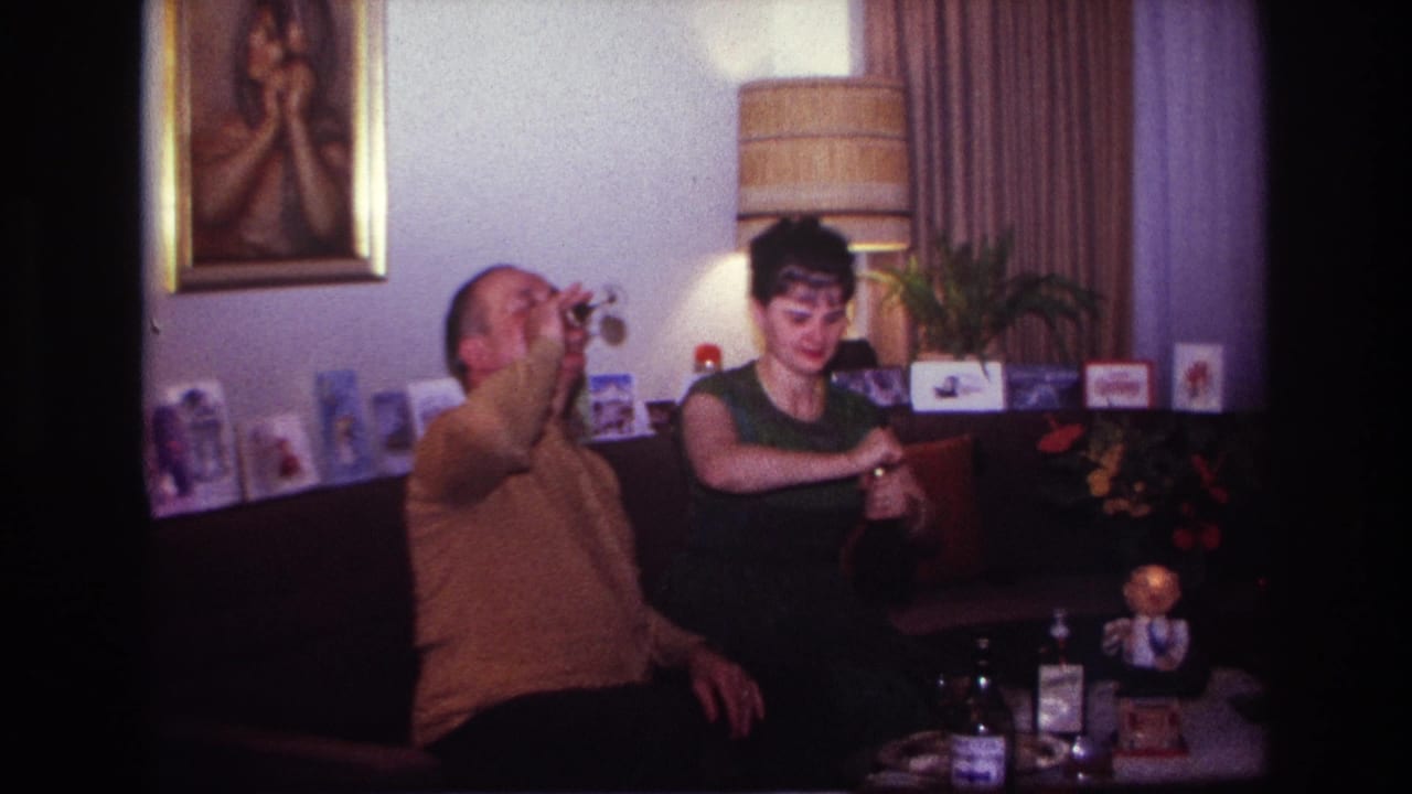1967 Rockaway Beach home-movie frame showing a seated man raising a drink while a woman sits beside him in a decorated living room.