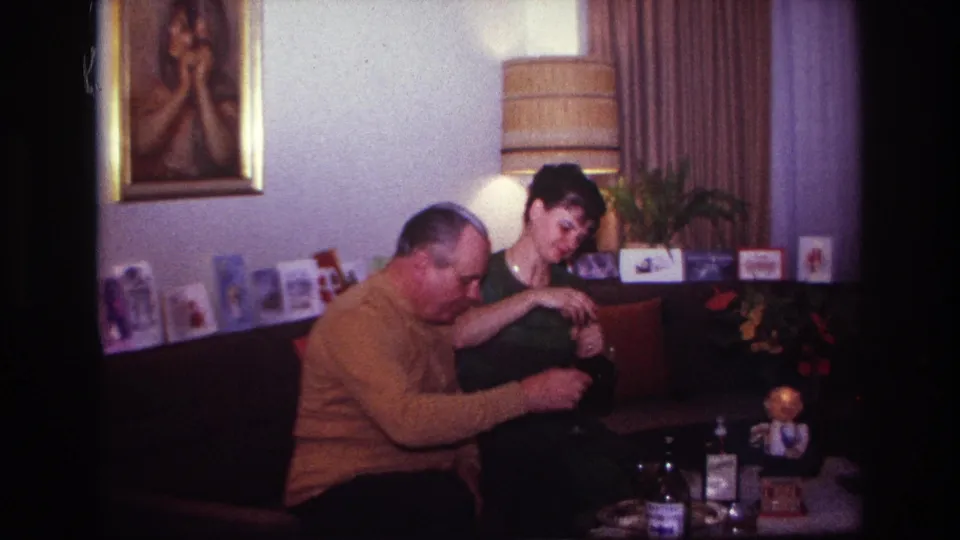 1967 Rockaway Beach interior frame with two adults seated close together, looking down as they handle drinks near a small table.