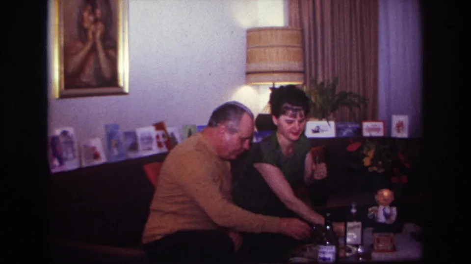 1967 Rockaway Beach home-movie frame of a woman reaching toward a table with bottles and glasses as a man leans forward beside her.