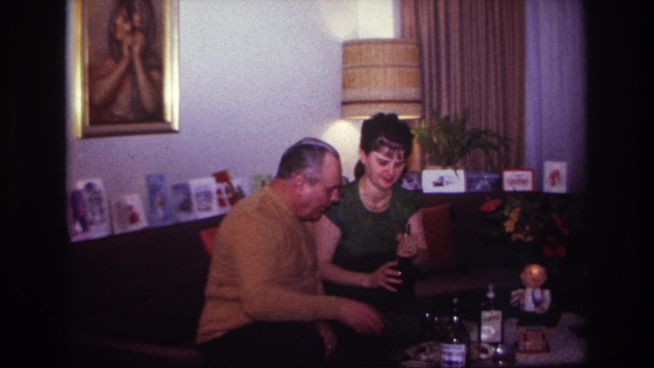 In a 1967 Rockaway Beach home-movie frame, a woman in a green dress sits beside a man in a mustard sweater near bottles and glasses.