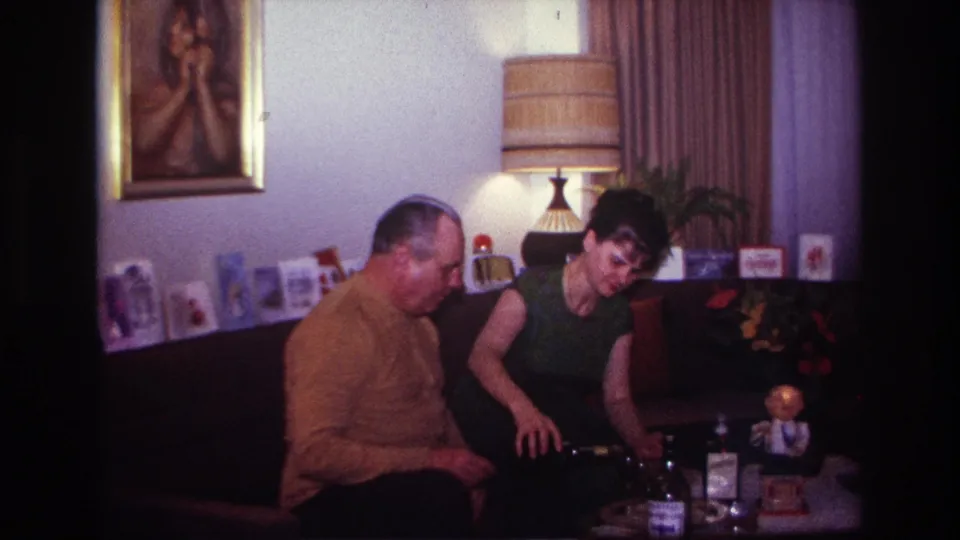 A woman in a green dress bends toward the coffee table with bottles and glasses in a softly lit 1967 domestic interior.