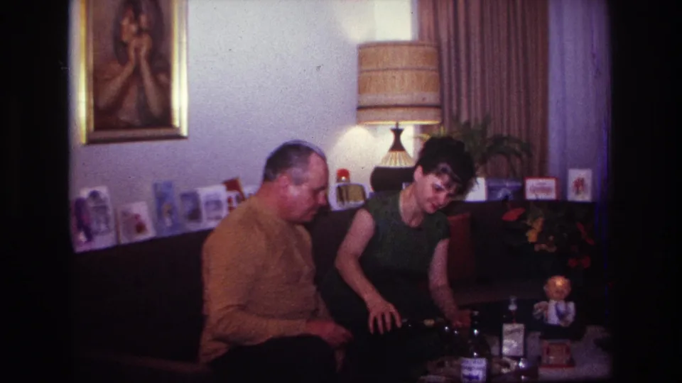 In a 1967 Rockaway Beach home interior, a man in a mustard sweater and a woman in a dark sleeveless dress sit near bottles and glasses.
