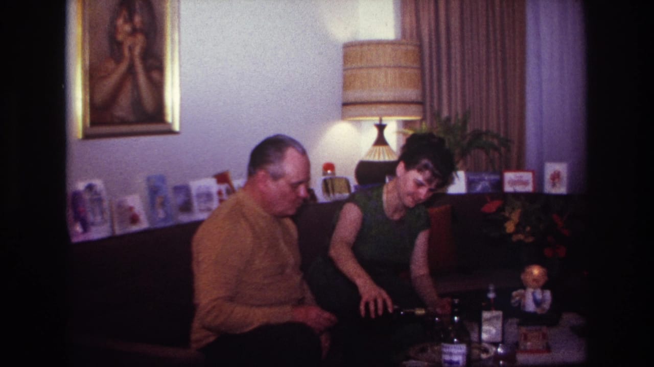 Woman leans toward bottles on a low table while the seated man looks down beside her.