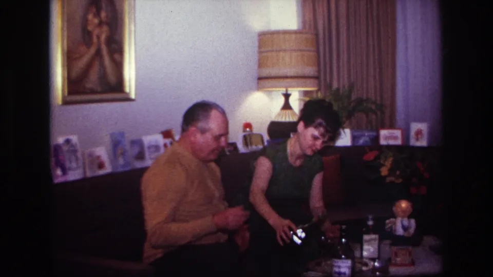 The man in a mustard sweater gestures while the woman works with a bottle or glass on the table in the 1967 Rockaway Beach interior.